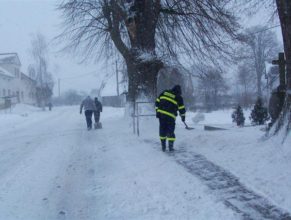 Dobrovolní hasiči odklízející sníh z chodníku u obecního úřadu.